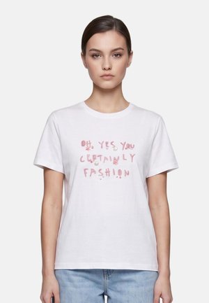 Jeune femme aux cheveux foncés attachés porte un t-shirt blanc avec un texte rose disant ""Oh, yes you certainly fashion"" et un jean bleu clair.