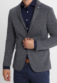 Uomo che indossa un blazer grigio texturizzato sopra una camicia button-up blu navy e pantaloni blu navy, che allaccia il blazer con la mano destra.