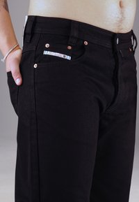 Schwarze Denim-Jeans im Slim-Fit mit kupferfarbenen Nieten, einem blau-weißen Etikett und einer Vordertasche.