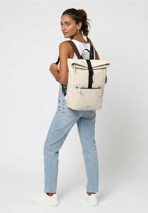 Femme portant un jean bleu clair et un débardeur blanc, avec un sac à dos beige avec des bretelles noires, debout devant un fond uni.