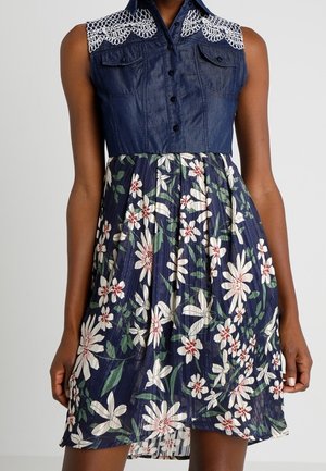 Robe sans manches avec haut boutonné en jean foncé et détail en dentelle blanche sur les épaules, associée à une jupe bleu marine avec imprimé floral blanc et rouge.