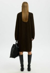 Přeceděný hnědý pletený svetr s dlouhými rukávy, doplněný nad kolena vysokými černými koženými botami a černou taškou přes rameno. Jednoduchý a strukturovaný design.