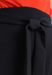 Námořnická látková páska s uzlíkem. Materiál vypadá hladce s mírnou strukturou. Součást oděvu s viditelným červeným vrškem pod ním.