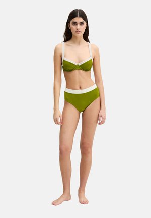 Młoda kobieta stojąca boso w zielono-białym bikini z wysokim stanem i regulowanymi ramiączkami na jednolitym białym tle.