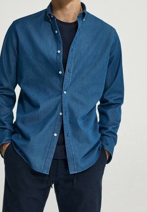 Mann trägt ein aufgeknöpftes blaues Jeanshemd über einem schwarzen T-Shirt mit den Händen in den dunklen Hosentaschen vor einfarbigem Hintergrund.