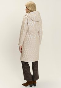 Donna con capelli biondi che indossa un cappotto imbottito beige con cappuccio, pantaloni a quadri scuri e stivali in suede marroni, in piedi e rivolta verso sinistra.