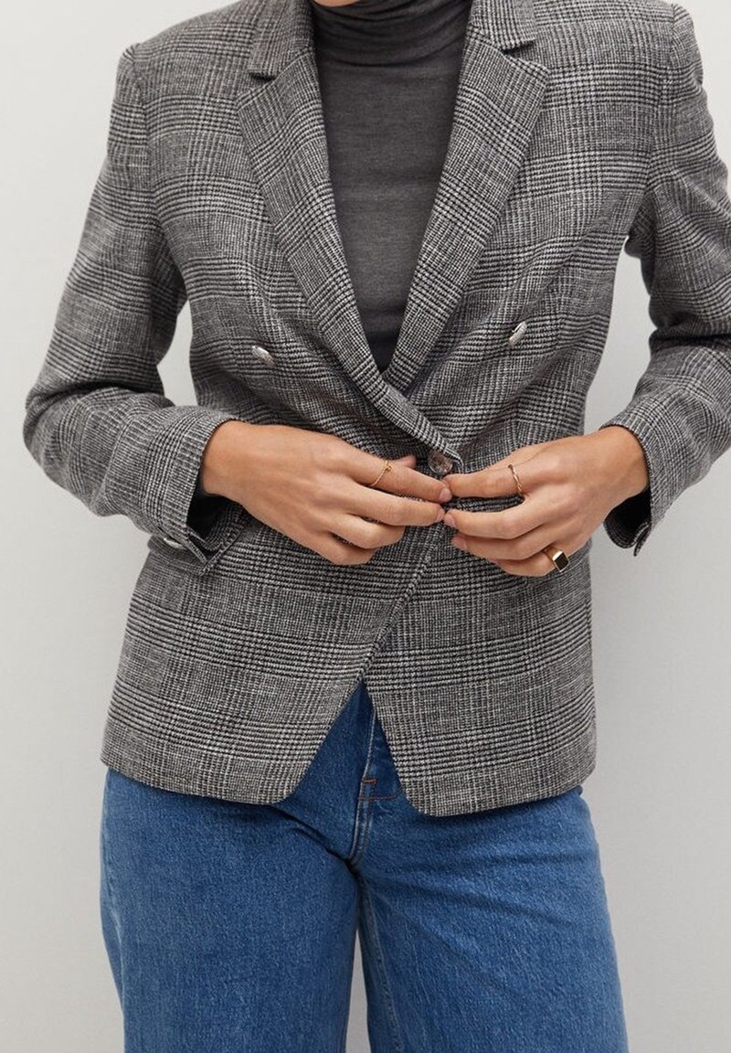 Grijze geruite blazer met een getailleerde snit, double-breasted ontwerp en metalen knopen, gecombineerd met een donker coltrui en blauwe spijkerbroek.