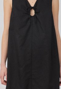 Robe noire sans manches avec un décolleté en V, détail en trou de serrure au centre, tissu texturé et lien à cordon au cou.