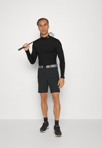Black long-sleeve shirt with logo, gray belt, and dark shorts. Model holds a golf club, wearing black sneakers and socks. Plain background.