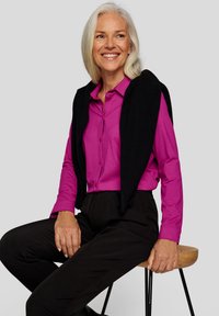 Magenta button-up shirt paired with black sweater draped over shoulders and black trousers; model seated on a wooden stool.