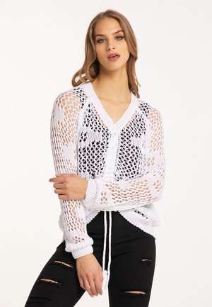 Jonge vrouw draagt een witte opengebreide trui over een zwart topje en gescheurde zwarte broek, poseert met gekruiste armen tegen een witte achtergrond.