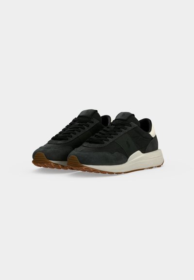 Ζευγάρι μαύρα sneakers με χαμηλό επάνω μέρος, καφέ λαστιχένιες σόλες, λευκές μεσαίες σόλες και μαύρα κορδόνια, εμφανίζεται σε απλό φωτεινό φόντο.