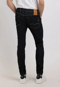 Jeans en denim foncé à coupe slim avec des surpiqûres dorées contrastantes, un patch en cuir à l'arrière et deux poches arrières courbées. Portés avec des baskets blanches.