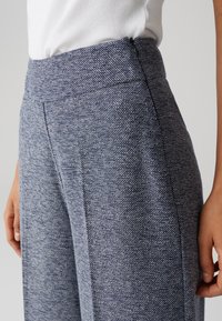 Pantalons larges en tissu bleu texturé avec un motif pied de poule subtil. Dotés d'une taille haute et d'une fermeture éclair sur le côté.