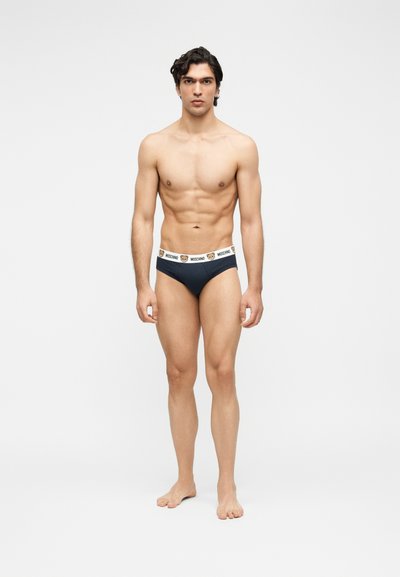 Νέος άνδρας στέκεται ξυπόλητος, φοράει σκούρα μπλε σλιπ με λευκή μέση που έχει πρόσωπα αρκουδάκια και το κείμενο «Moschino», σε απλό φόντο.