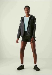 Joven mujer de pie vistiendo una chaqueta cortavientos negra, una camiseta azul clara, pantalones cortos negros, calcetines blancos y zapatillas negras contra un fondo liso.