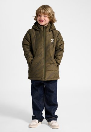 Lachend kind met krullend blond haar, gekleed in een olijfgroene gewatteerde jas, donkerblauwe spijkerbroek en witte sneakers, tegen een effen achtergrond.