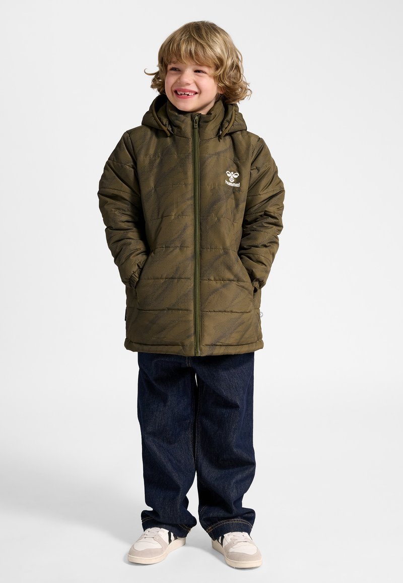 Uśmiechnięte dziecko o kręconych blond włosach, ubrane w oliwkowo-zieloną pikowaną kurtkę, ciemnoniebieskie dżinsy i białe trampki na jednolitym tle.