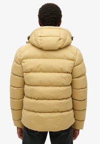 Chaqueta de plumas beige con capucha, con costuras horizontales, un acolchado prominente y un logo "SUPERDRY" en la parte trasera. Material texturizado.