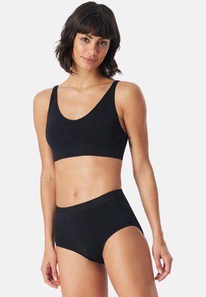 Kvinde med kort mørkt hår iført sort ærmeløs crop top og sort højtaljet undertøj mod en ensfarvet hvid baggrund.