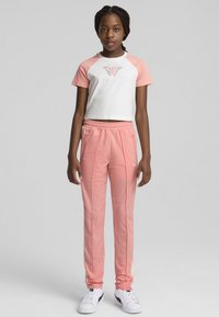 Bela crop majica z rožnatimi rokavi in grafiko metulja, v kombinaciji s svetloroznimi trenirkami z bočnimi žepi. Bela športna obutev dopolnjuje outfit.