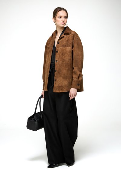 Nő barna gombos dzsekit visel fekete széles szárú nadrág fölött, fekete kézitáskát tart, és egy egyszerű fehér háttér előtt áll.