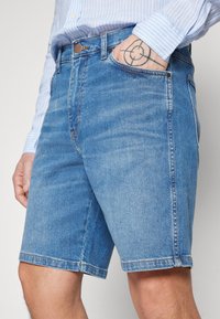 Shorts de denim en azul claro con corte recto, diseño de cinco bolsillos y costuras en contraste. La tela muestra un ligero desgaste y textura.