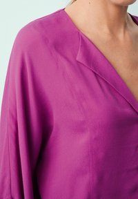 Blusa fucsia con scollatura a V, vestibilità comoda e tessuto leggero e testurizzato. Presenta maniche ampie e una lucentezza sottile.