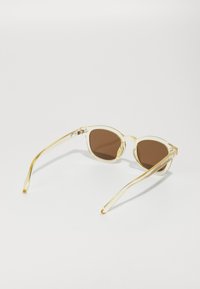 Ochelari de soare clar, de formă rotundă, cu lentile maro; ramă transparentă galbenă; design elegant; brațe subțiri cu o ușoară curbă; branding minim pe tâmple.