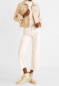 Persoon in een beige cropped jack, een witte button-up shirt, hoge witte wijde jeans en beige high-top sneakers.