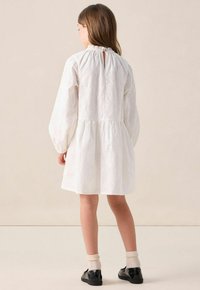 Fille se tenant de dos, portant une robe blanche à manches longues avec des broderies florales, des chaussettes crèmes et des chaussures noires sur un fond beige.