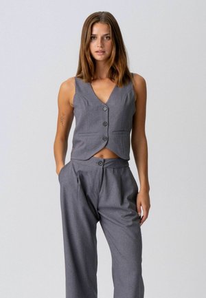 Kobieta o brązowych włosach do ramion, ubrana w szarą, bezrękawnikową kamizelkę zapinaną na guziki oraz pasujące, wysokowodne, szerokie spodnie.