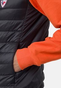 Černá péřová vesta s horizontálním prošíváním a boční kapsou na zip, vrstvená přes oranžový pletený svetr s ribbenými manžetami. Logo viditelné na vestě.