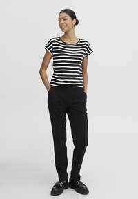 Top de rayas en blanco y negro de manga corta combinado con pantalones negros ajustados y zapatos negros gruesos. El modelo se encuentra de pie con las manos en los bolsillos.