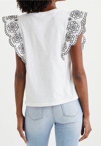 Vit bomullströja med svart blommönsterbroderi på volangärmar, med en avslappnad passform och rundad nederkant, tillsammans med ljusblå jeans.