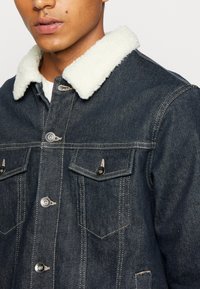 Giacca di jeans scuri con colletto in ecopelliccia bianca, due tasche sul petto e chiusure a pulsante con dettagli in metallo. Dettagli di cucitura distintivi in vista.