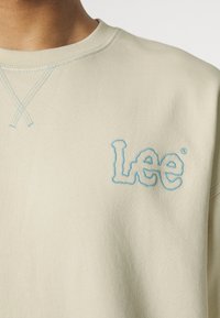 Světle béžová mikina vyrobená z bavlny, vybavená modrou vyšívanou značkou "Lee" na přední straně a křížkovým stehem kolem výstřihu.