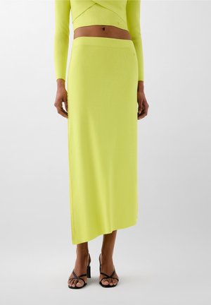 Femme portant une jupe midi asymétrique à taille haute jaune et un haut court assorti à manches longues, assortis à des sandales à talons noires à brides.