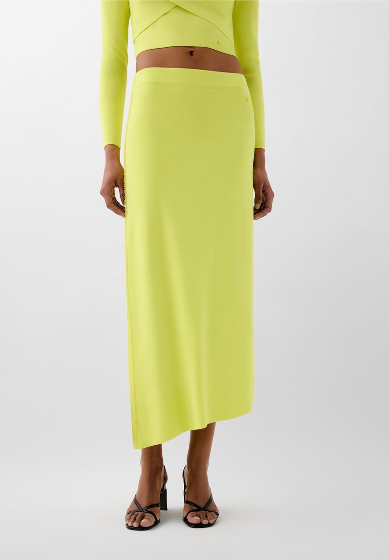 Femme portant une jupe midi asymétrique à taille haute jaune et un haut court assorti à manches longues, assortis à des sandales à talons noires à brides.