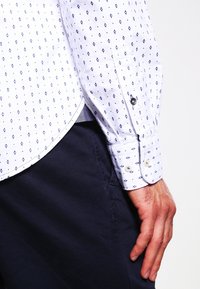 Weißes Hemd mit navyfarbener, geometrischer Rautenstruktur, mit Knopfmanschetten und gebogenem Saum. Getragen mit dunklen Hosen, die den Stoff und die Details zur Geltung bringen.