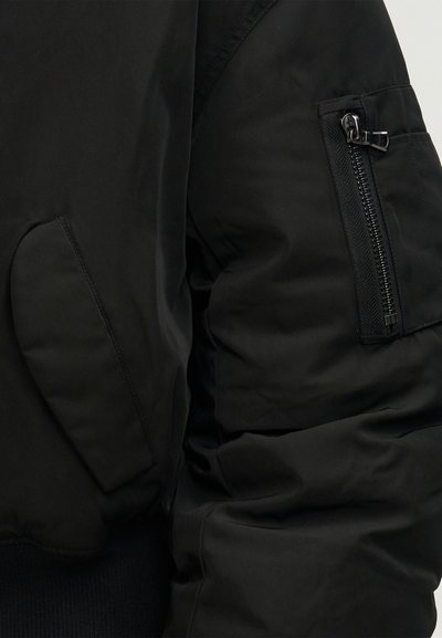 Μανίκι μαύρου μπουφάν με τσέπη με φερμουάρ και λοξή τσέπη με καπάκι, δείχνοντας υφή και λεπτομέρειες ραφής.