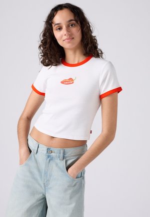 Νεαρή γυναίκα με σγουρά μαλλιά φοράει λευκό κοντό μπλουζάκι με κόκκινο περίγραμμα και λογότυπο με τσίλι, σε συνδυασμό με ανοιχτά μπλε τζιν ψηλόμεσα.