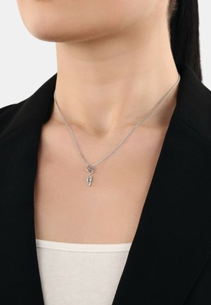 Zilveren ketting met een kleine schoen charm, gedragen boven een lichtgekleurde top, tegen een achtergrond van een donkere blazer.