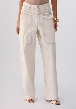 Donna che indossa pantaloni crema a gamba larga e vita alta con grandi tasche applicate frontali e laterali, abbinati a sandali open-toe e braccialetti dorati.