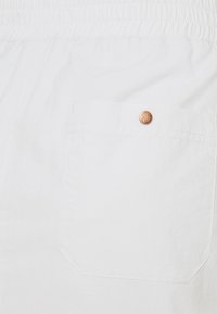 Pantalones cortos de algodón blancos con una cintura elástica, que cuentan con un bolsillo trasero con un botón de cobre. Textura suave con un diseño minimalista.
