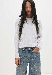 Pulover tricotat de culoare gri deschis, cu mâneci lungi, gât rotund și tiv cu nervuri, purtat cu blugi evazați de denim albastru, cu un aspect uzat.