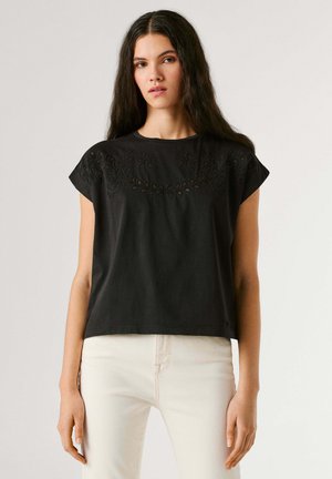 Jonge vrouw met lang donker haar, gekleed in een zwart geborduurd topje met korte mouwen en witte broek, tegen een effen achtergrond.