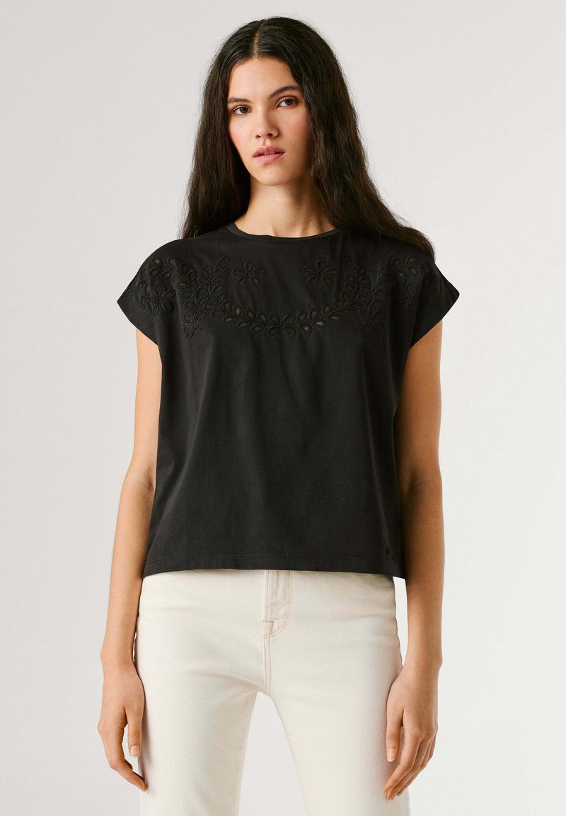 Mlada ženska z dolgimi temnimi lasmi, oblečena v črno vezeno majico s kratkimi rokavi in bele hlače, stoji pred enobarvnim ozadjem.