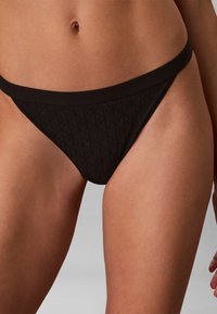 Bas de bikini noir taille haute avec un motif matelassé subtil porté sur la partie basse du torse et le haut des cuisses d'une personne sur un fond uni.