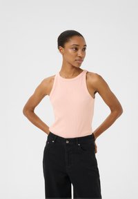 Lichtroze geribde tanktop met een hoge halslijn en logo-detail, gecombineerd met hoge zwarte spijkerbroeken, staande tegen een neutrale achtergrond.